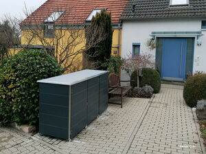 . Ein modernes Haus mit Vorgarten, Bank, blauen Türen, Pflanzen und Abfallbehälterboxen auf gepflastertem Weg.