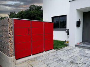 . Roter Paketkasten vor einem modernen Haus, mit Holzlager links und Terrasse im Vordergrund.