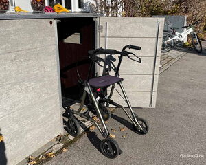 . Ein Rollator steht vor einem offenen Holzschrank auf einem Weg, daneben sind zwei Fahrräder.