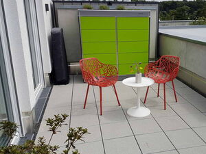 . Zwei rote Stühle, ein kleiner weißer Tisch mit Vase, grüne Wand auf einer Terrasse.