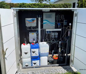 Ein Technikschrank mit Pumpen, Schläuchen und Chemikalienkanistern für Pool oder Wasseraufbereitung.