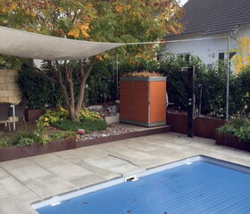The image depicts a backyard scene which includes:

1. A swimming pool with a blue cover.
2. A paved area around the pool.
3. A garden with various plants and a tree with autumn foliage.
4. A rectangular structure with an orange casing, possibly a storage unit or utility box.
5. A sunshade canopy.
6. A building or house with a visible part of the roof in the background.