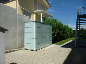 . The image shows a rectangular, modern storage unit or cabinet placed outside a building. It is set on a paved area with stairs and greenery nearby. The cabinet has a sleek design with horizontal lines, possibly indicating separate compartments or panels.