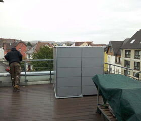 Ein Mann lehnt sich auf einem Balkon über das Geländer, neben einem großen, grauen Schrank. Sicht auf Häuser im Hintergrund.