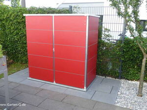 . Ein rotes Gartenhaus mit Metallrahmen auf einer gepflasterten Fläche, umgeben von Pflanzen und Bäumen.