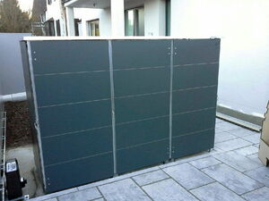 . The image shows a gray metal storage cabinet or a shed placed outdoors on a paved surface. It's positioned against a building, and has a flat top and handles for opening. The setting appears to be a patio or backyard.

