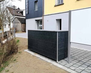 Ein modernes Gebäude mit dunklem Zaun, Mauerwerk, Pflanzen und Gehweg. Häuser und Bäume im Hintergrund.