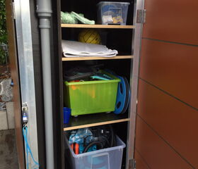 Ein Schrank mit Regalen: Plastikboxen, Tücher, grüne Box, gelber Ball, Schlauch, Poolzubehör.