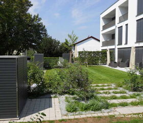 . Moderner Garten mit Rasen, Büschen, gepflasterten Wegen, zwei Schuppen, Terrasse mit Möbeln und Mehrfamilienhaus.
