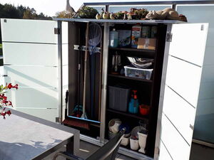 . Ein Gartenschrank auf einem Balkon mit Gartengeräten, Reinigungsmitteln und Pflanzen oben.
