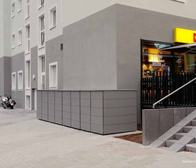 . Außenansicht eines Gebäudes mit einer Fahrradabstellanlage, Treppe und Eingang zu einem Geschäft namens "Der B.".