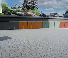 Kleine Schränke an einer Wand, bunte Türen, Pflastersteine, Autos auf erhöhtem Parkplatz im Hintergrund.