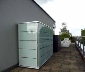. The image shows a rooftop or terrace area. There is a frosted glass storage cabinet with metal accents against a wall. Near it are large white bags and an object that appears to be a black structure. The area has a paved surface and a metal railing along the edge. Some trees and a distant cityscape or landscape are visible beyond the railing.