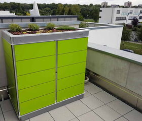 Ein grüner Schrank auf einer Terrasse, mit einem bepflanzten Dach im Vordergrund, umgeben von modernen Gebäuden.