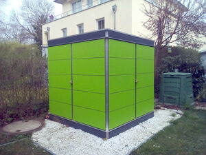 . The image shows a green structure in a garden setting. The structure appears to be a shed or a small building with a modern design, featuring smooth, bright green panels. It is surrounded by white gravel and the background includes a residential building, trees, and another green object that resembles a compost bin or storage unit.