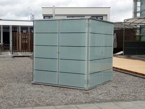 . This image shows a glass and metal structure with a modern design. It is located on a gravel and paved area, possibly an outdoor space adjacent to buildings. The structure has opaque glass panels and is surrounded by other architectural features, including a wooden deck and additional buildings.