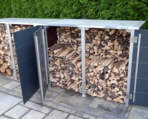 . The image shows a storage unit for firewood. It has metallic doors, some of which are open to reveal neatly stacked wood inside. The storage is positioned on a patio, and there is greenery in the background.

