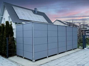 . Ein grauer Geräteschuppen steht vor einem Haus mit Solarpanelen auf dem Dach.