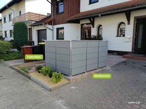 . Fahrräder und Mülltonnen sind vor einem Haus in einem grauen Gehäuse aufbewahrt.