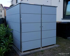 The image shows a modern, metal storage shed with a sleek, panel design. It's situated outdoors, next to a building, with some greenery on the left side. There are two doors in the front, and it's placed on a paved area.