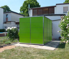 . The image shows a green cube-shaped structure made of panels situated in a garden area. It's placed on a paved surface and surrounded by grass, flowers, and bushes. There are houses in the background under a clear sky.