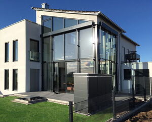 . The image depicts a modern house with a sleek, contemporary design. It features large glass windows and a mix of white and dark structural elements. The house is set on a green lawn with a paved area and is enclosed by a simple fence. The sky in the background is clear with some clouds, indicating a sunny day.