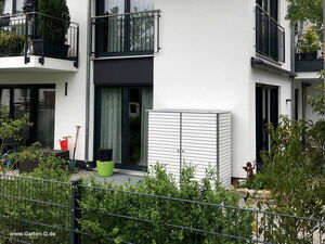 . The image shows the exterior of a modern residential building. There is a patio area with a white storage cabinet, a green plant pot, and some gardening tools. The building has a balcony with plants and railings. There are also decorative elements in the garden. A metal fence separates the garden from the foreground.
