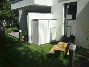. The image contains the following elements in an array:

1. A white building with a modern design.
2. A small white shed or storage box with a slatted design.
3. A wooden table with two benches, likely for outdoor seating.
4. Green grass covering the ground.
5. A deck or patio area with potted plants.
6. Two rectangular outdoor light posts.
7. A doorway with glass and a reflection in it.

This outdoor area appears to be part of a backyard or garden setup adjacent to a modern home.