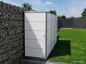 . Ein moderner, weißer Gartenschrank steht an einer Gabionenwand auf einer Rasenfläche.