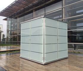 . The image shows a modern-looking building or structure with a prominent cube-shaped object in the foreground. The cube is made of frosted or translucent glass with horizontal lines. It is positioned outside on a wet, polished surface, possibly a patio, adjacent to a building with glass and metal elements. The style is sleek and contemporary.