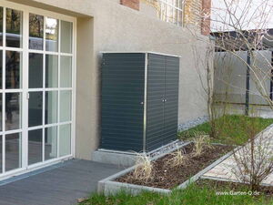 . The image shows a small garden shed or storage unit positioned against the exterior wall of a building. It is made of dark-colored panels and has a flat top. To the left, there is a large window or glass door. In the foreground, there's a small landscaped area with plants and paved walkways.