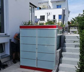 . The image depicts a modern, sleek outdoor storage unit placed next to a house. It features a minimalist design with frosted glass panels and a red accent at the top. There are stairs on the right side, and a person is partially visible on the left. The environment is clean and well-maintained, suggesting a residential area.