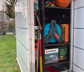 . This image shows the contents of an outdoor storage cabinet. Inside, you can see:

1. Various gardening tools and equipment.
2. A basketball and some brightly colored juggling balls.
3. A hose reel.
4. Boxes labeled "Gardena" with gardening supplies.
5. A bag of grill charcoal briquettes.
6. Other items like a saw and small boxes, possibly for storage or organization.
