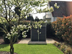 . The image depicts a garden scene with the following elements:

1. A tree with white blossoms in the foreground.
2. A neatly manicured lawn.
3. A dark-colored tall structure, possibly a solar panel or light fixture, on a small gravel area.
4. A house with a tiled roof in the background.
5. Various shrubs and plants surrounding the area.
6. A small garden spotlight in the lower left corner.