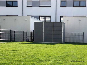 . The image shows a modern outdoor storage unit or shed with a sleek design, situated on a grassy area. There's a metal fence around the area, and the background features a building with multiple windows.