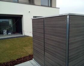 . The image shows a backyard or garden area. There's a well-maintained grass lawn with a mulched edge. On the right side, there's a wooden or wood-paneled structure, possibly a shed or storage unit. In the background, there is a modern building with glass doors leading to a patio. On the patio, there are some items stored in plastic containers. The setting appears to be organized and well-kept.
