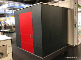 . The image shows a large, box-like structure with a modern design. It's primarily dark with a contrasting red door featuring metal handles. The environment looks like a display area or exhibition, indicated by the surrounding booths and banners. The text on the structure and background suggests it's part of a trade show or exhibition display.