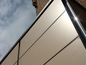. The image shows a modern building structure with sleek, smooth panels contrasting against an older brick building with detailed architecture. The photo is taken from an upward angle that emphasizes the contrast between the two architectural styles. The sky is partly cloudy.