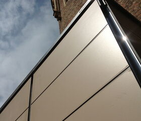 . The image shows a modern building structure with sleek, smooth panels contrasting against an older brick building with detailed architecture. The photo is taken from an upward angle that emphasizes the contrast between the two architectural styles. The sky is partly cloudy.
