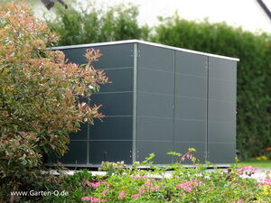 . The image shows a modern, dark-colored garden shed. It is situated in a garden surrounded by various plants and foliage. The shed has a sleek design and appears to be made of metal panels. The surroundings include green shrubs and flowering plants.
