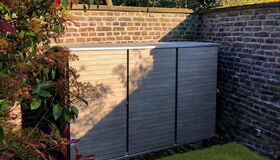 . The image shows an outdoor storage unit placed against a brick wall. It is surrounded by greenery, with plants on one side and grass at the base. The storage unit has a modern, wooden design with multiple vertical panels.