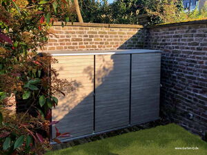 . The image shows an outdoor storage unit placed against a brick wall. It is surrounded by greenery, with plants on one side and grass at the base. The storage unit has a modern, wooden design with multiple vertical panels.