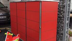 . The image shows a red storage unit or cabinet next to a wall made of gabion baskets filled with rocks. In front of the cabinet, there is a small playground spring rider shaped like a red and green horse. It appears to be located in an outdoor area, possibly near a residential or parking space.