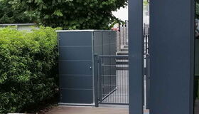 . The image shows an outdoor scene featuring a modern storage unit or cabinet with a sleek, dark-colored design. It's positioned near a metal gate and surrounded by greenery, including bushes and trees. There is also a wooden deck area visible in the foreground.