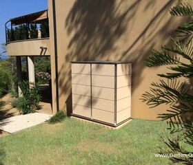 . The image shows an outdoor scene featuring a modern building with a rounded balcony and a set of stairs. There is a rectangular structure or cabinet against the wall, and the area is surrounded by greenery, including various plants and trees. The grass and shadow on the wall suggest it is a sunny day.