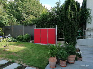 . Ein Garten mit rotem Schrank, Pflanzen, Rasen und Gehwegplatten. Im Hintergrund sind Bäume und eine Hecke.