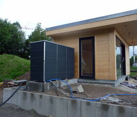 . The image depicts a modern, small, wooden building with large windows. Near the building, there is a large, rectangular, black box-like structure with cables connected to it, which might be part of a heating or cooling system. The area around the building is under construction and includes some construction materials and debris, along with a grassy area and trees in the background.