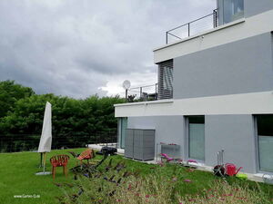 . Modernes Haus mit Terrasse, Gartenstühle, Schirm und Gartenwerkzeuge im grünen Garten, bewölkter Himmel.