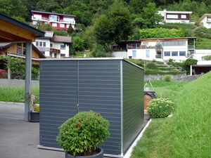 . The image shows a modern garden shed or storage unit with a metal exterior. It is situated on a paved area with a planted shrub in a pot nearby. In the background, there are several houses on a hillside, surrounded by greenery and trees.