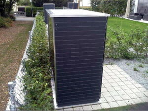 . The image shows a row of tall, rectangular cabinets or enclosures located outdoors. They appear to be situated on a paved area next to a garden or lawn, with greenery and a metal fence nearby. The enclosures have a dark finish with horizontal lines and a flat top. They seem to be neatly arranged in a line extending into the background.